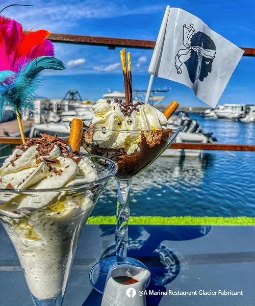 Deux coupes de glace avec chantilly et un drapeau corse planté dedans, au restaurant A Marina en Corse, avec vue sur les bateaux du port de plaisance de Sari-Solenzara